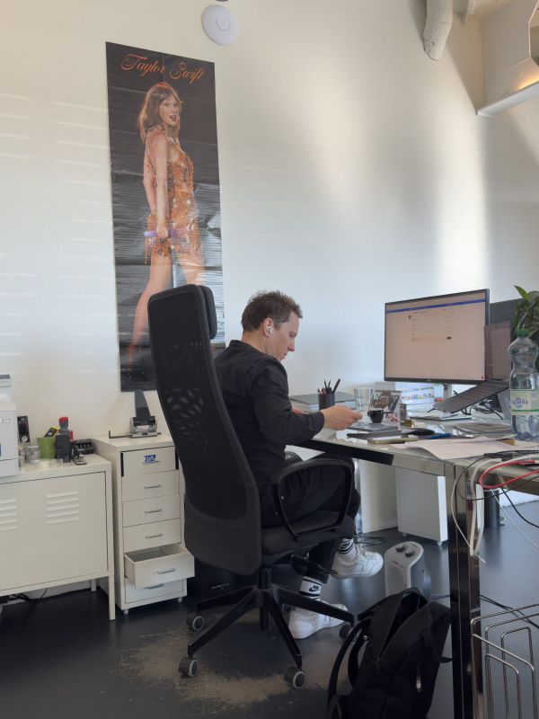 Raphael sitzt an seinem Arbeitsplatz, im Hintergrund ist ein riesiges Taylor Swift Plakat zu sehen.
